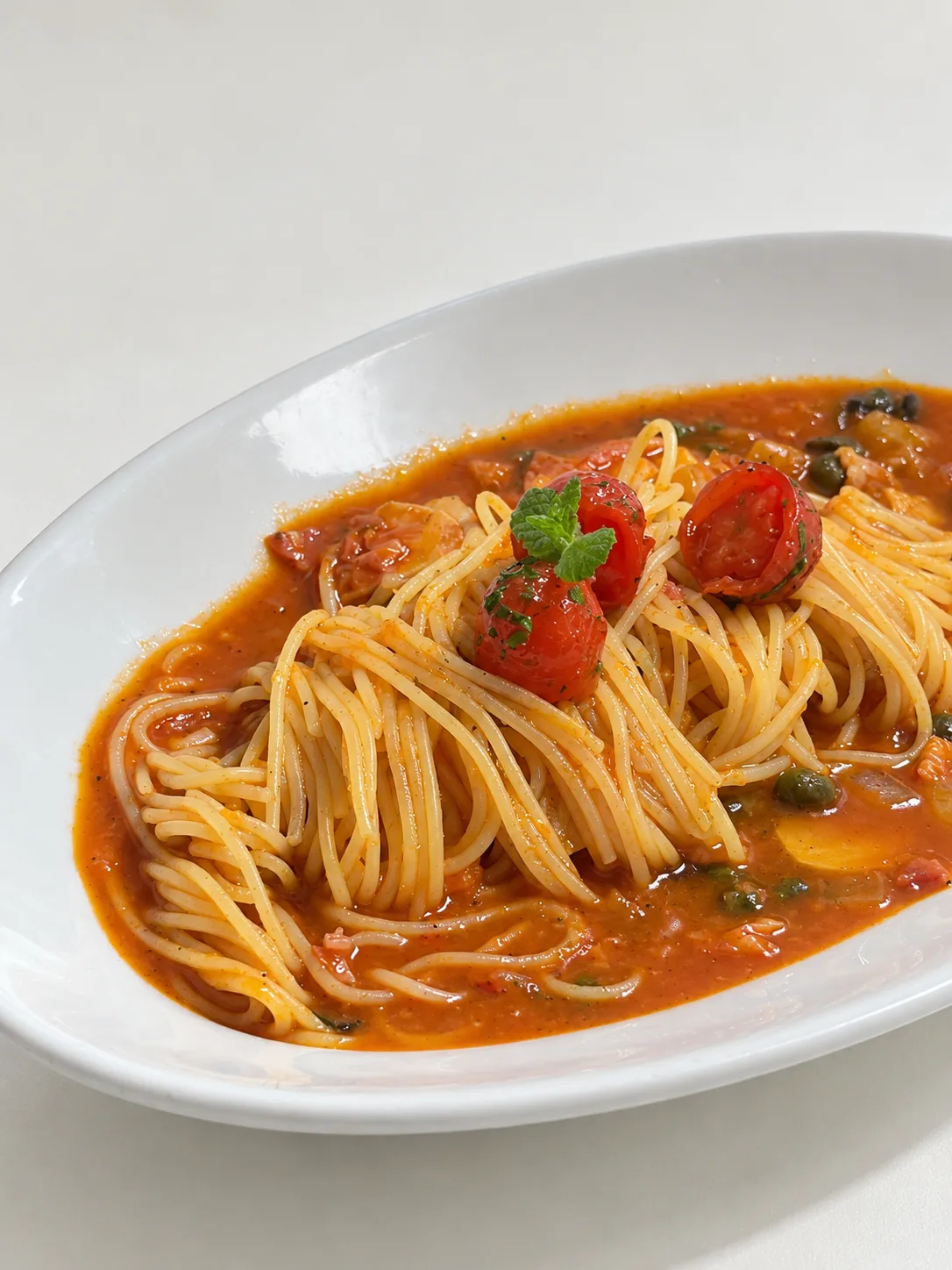 흰 타원 접시의 토마토소스 파스타 — 케이퍼, 올리브, 방울토마토, 민트 잎