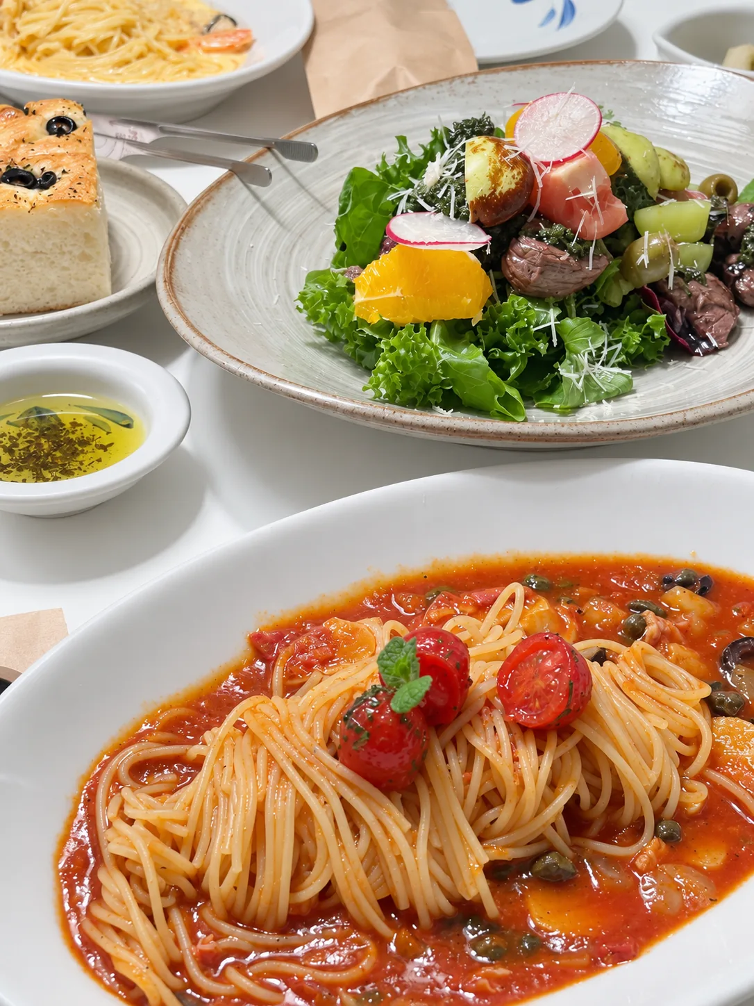 한 상 — 앞쪽 흰 접시의 토마토소스 파스타(케이퍼·올리브·방울토마토·민트), 뒤쪽 회색 도자기 접시의 샐러드(고기 조각·오렌지·래디시·올리브), 올리브를 올린 빵, 허브를 띄운 올리브유